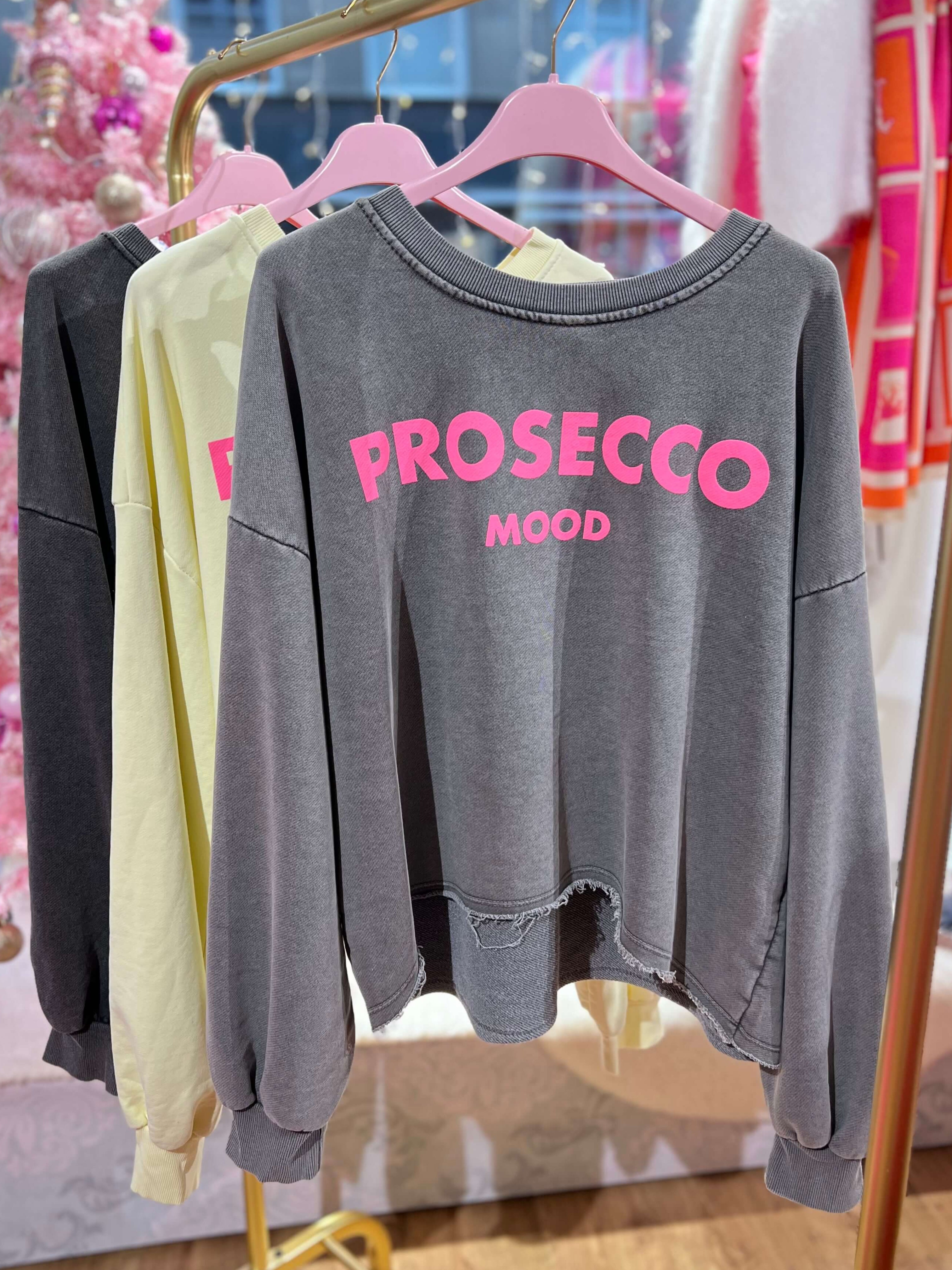 Grauer Frühlingssweater aus Baumwollgemisch im Used-Look mit Aufdruck „Prosecco Mood“, locker geschnitten, mit angesetztem Saum und langen Ärmeln, hängend an einem Kleiderbügel.