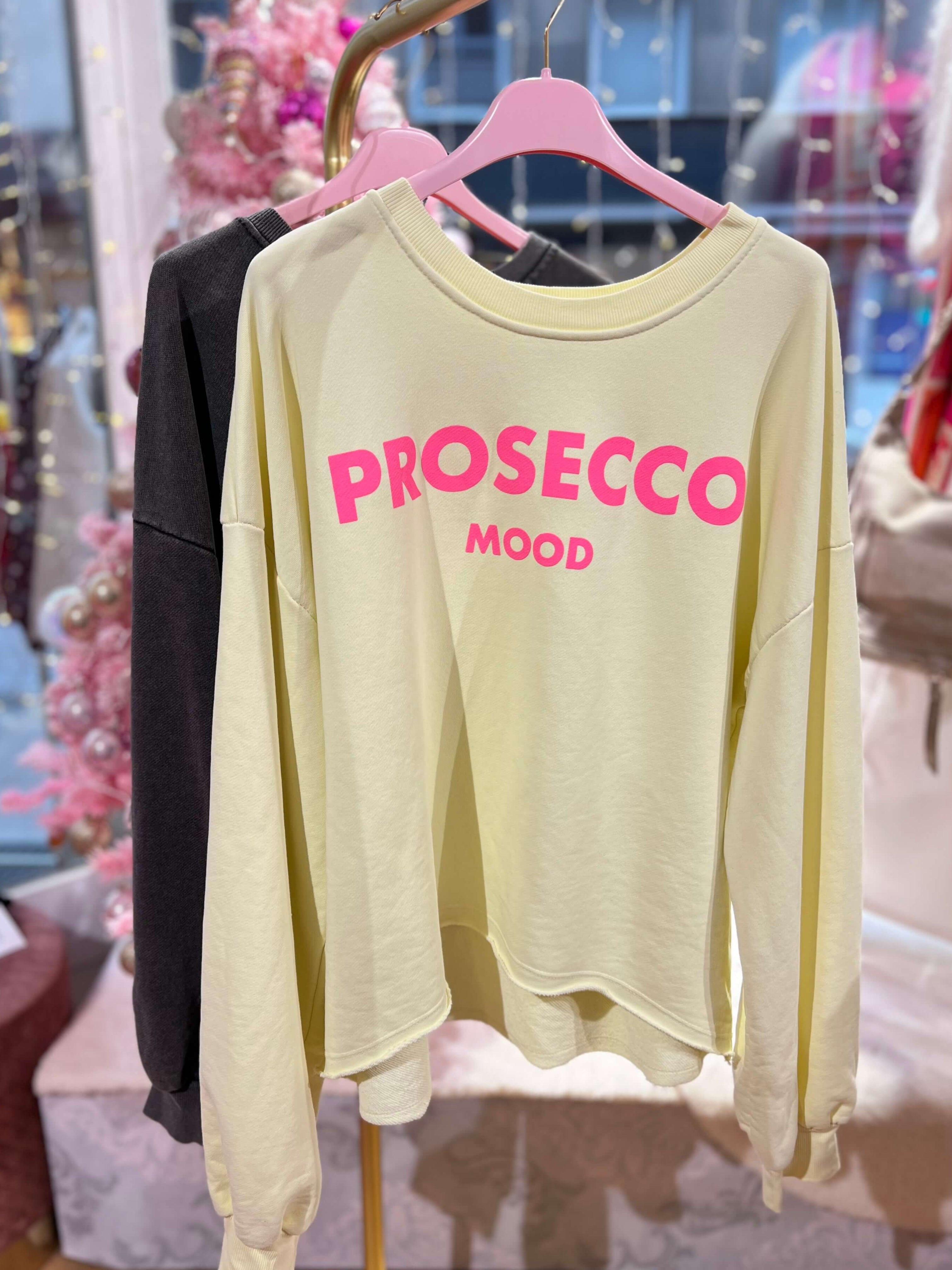 Gelber Frühlingssweater aus Baumwollgemisch im Used-Look mit Aufdruck „Prosecco Mood“, locker geschnitten, mit angesetztem Saum und langen Ärmeln, hängend an einem Kleiderbügel.