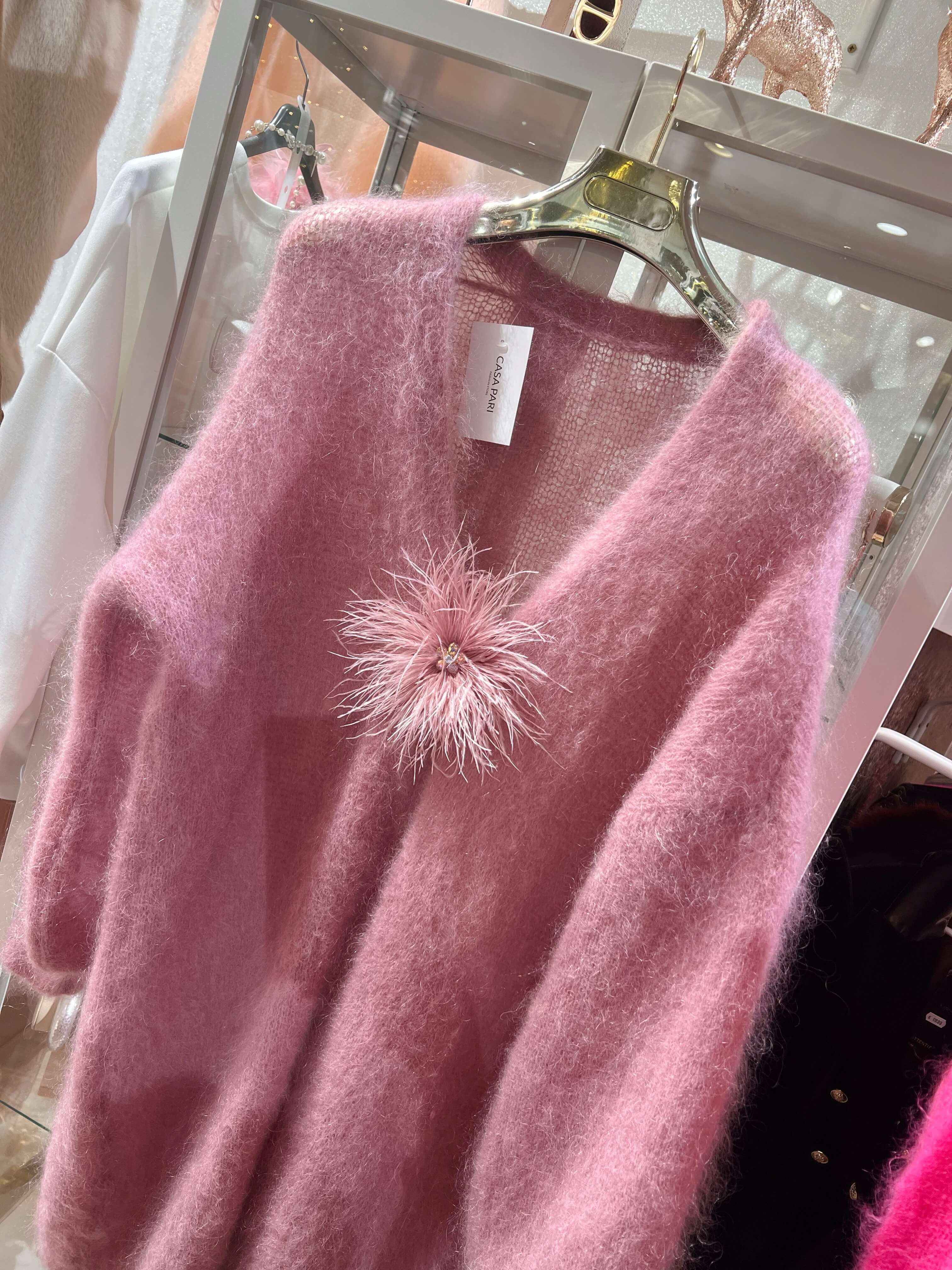 Rosa Baby-Alpaka-Pullover mit V-Ausschnitt auf Kleiderbügel, vorne mit einer rosa Federbrosche dekoriert, im Boutique-Innenraum fotografiert.