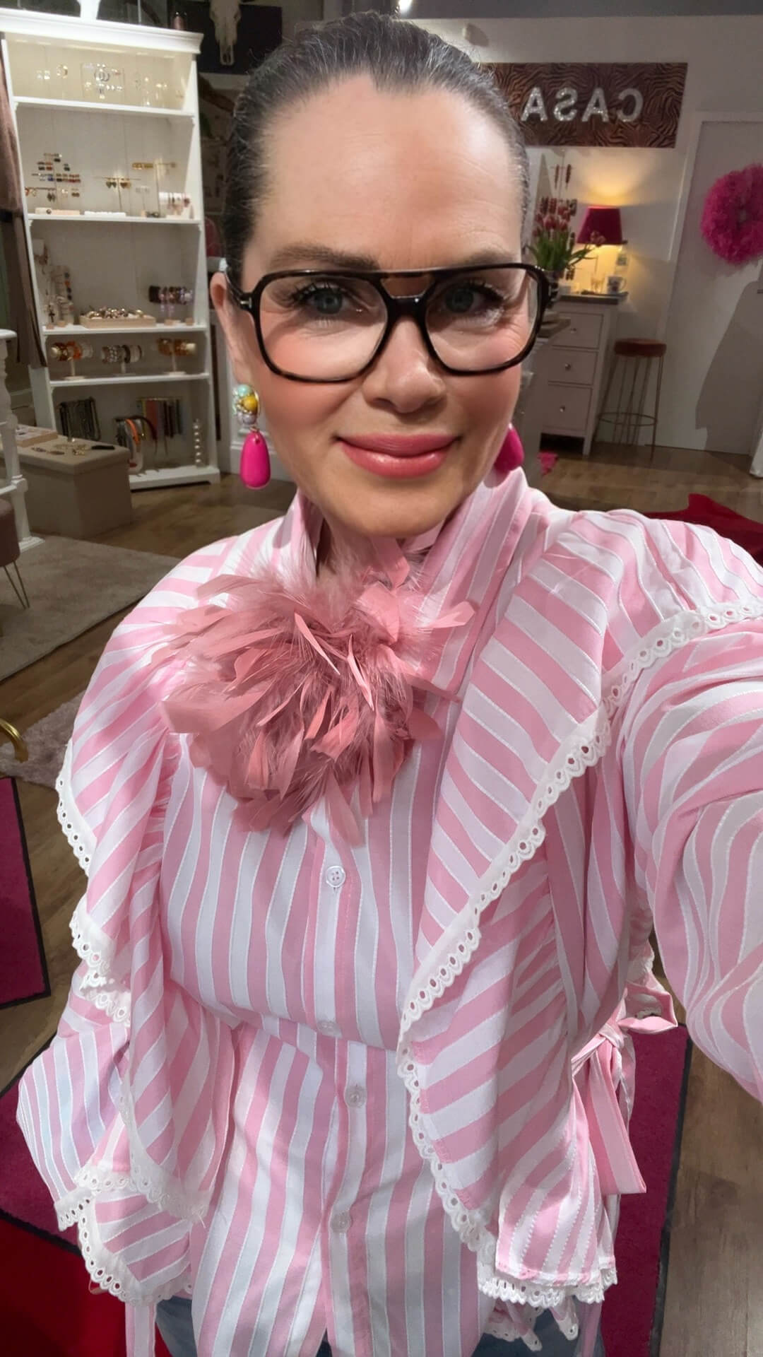 Person mit dunkler Brille trägt eine rosé-weiß gestreifte Bluse mit Spitzenborten und Schleifendetail am Kragen, dazu pinke Ohrringe, aufgenommen in einem Modegeschäft.