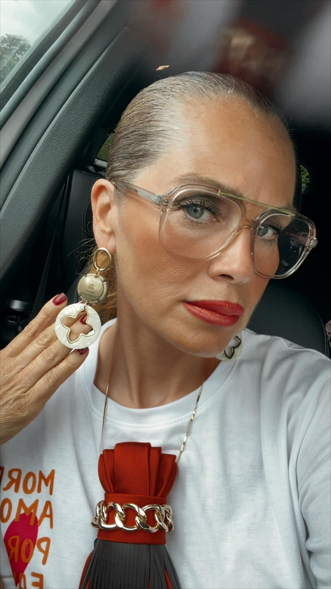 Frau trägt goldene, lange Ohrringe sie sitzt in einem Auto hat ein weißes Shirt an eine große Brille. Trägt eine Krawatte