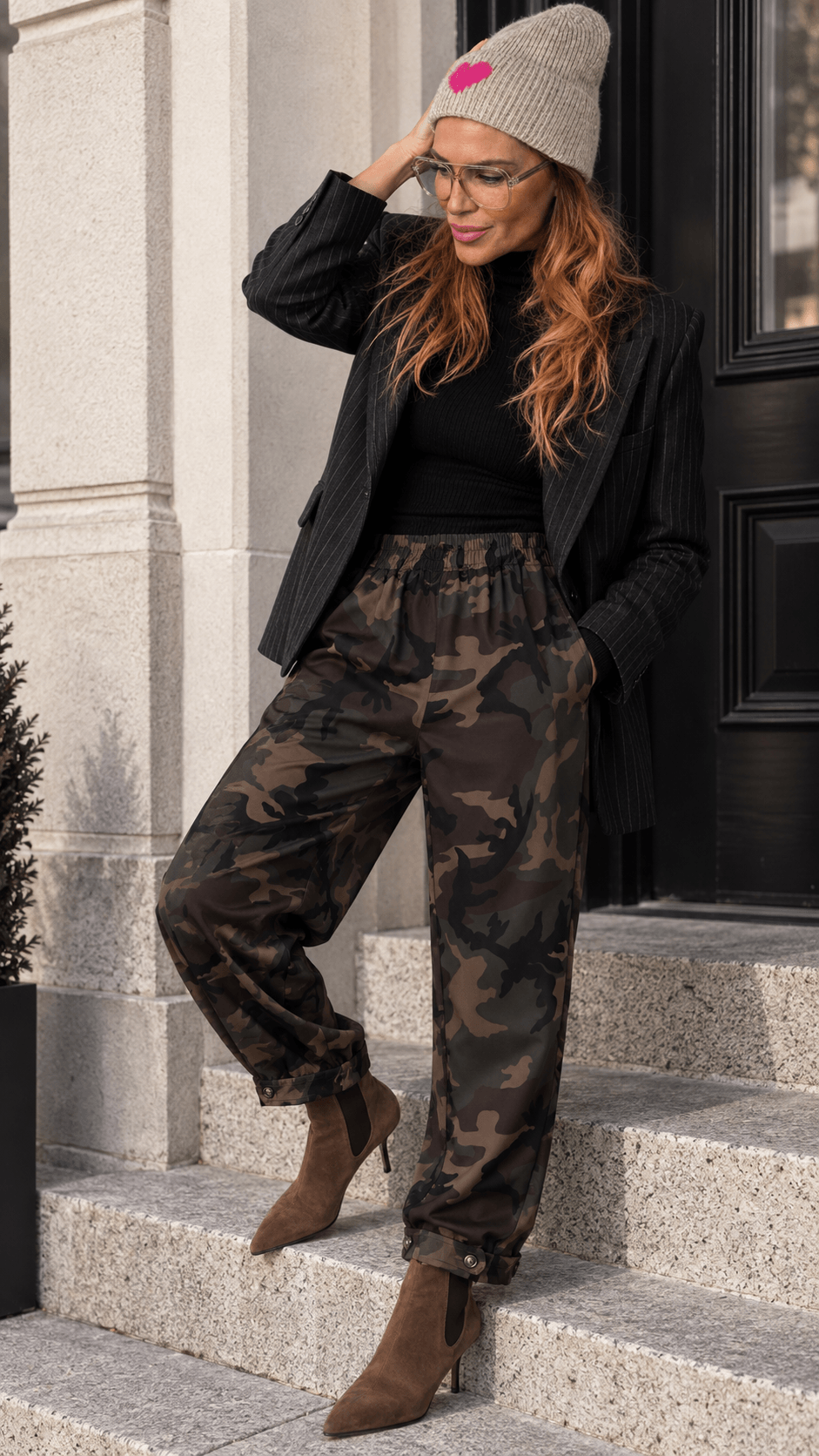 Frau trägt die Barrel Pants Urban Camo mit schwarzem Blazer, braunen Stiefeletten und beiger Mütze auf einer Steintreppe.