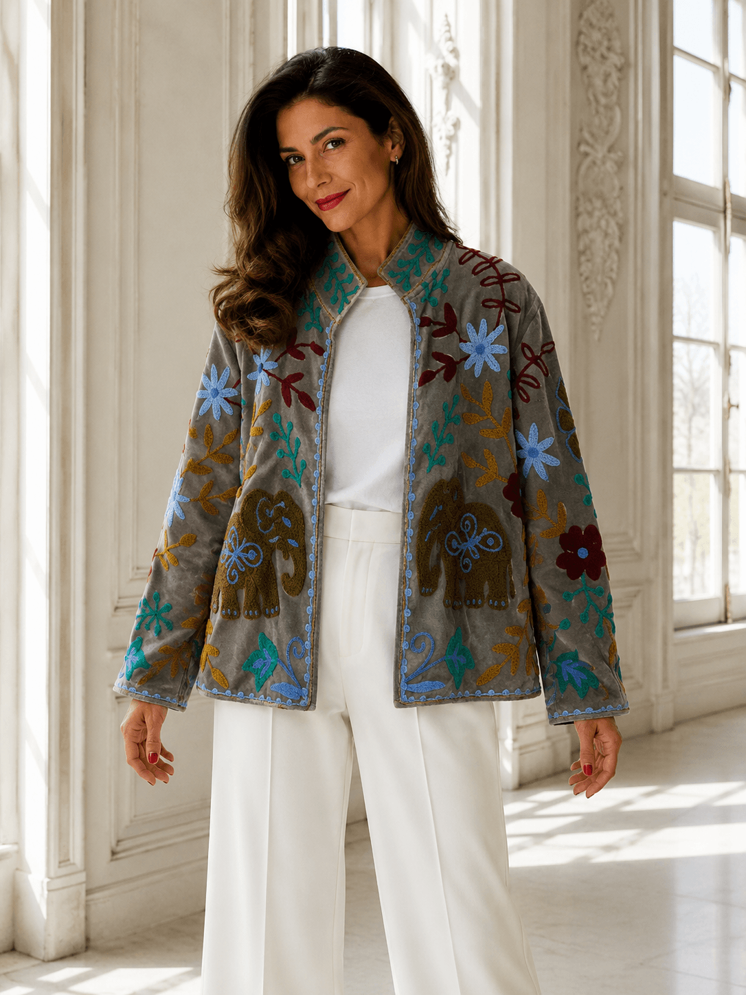 Frau trägt eine bestickte Jacke mit bunten floralen Stickereien und Elefantenmotiven in einem hellen eleganten Innenraum, Frontalansicht.