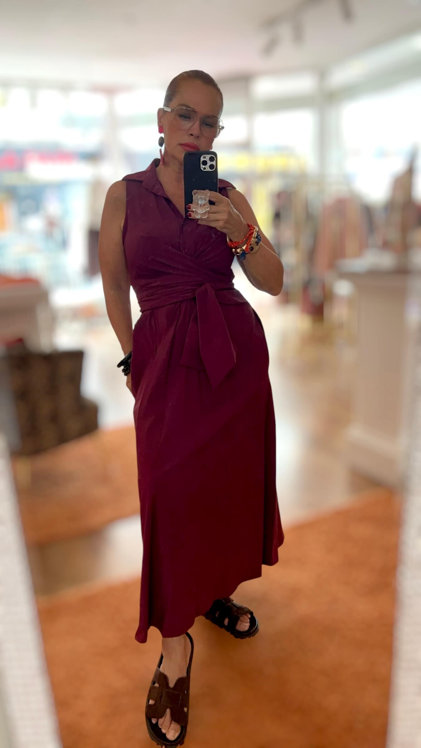 Frau trägt ein bordeauxrotes Wickelkleid mit Bindegürtel und Knopfleiste. Das ärmellose Kleid wird in der Taille gebunden und fällt fließend bis zum Knöchel. Kombiniert mit einer dekorativen Brosche am Ausschnitt und farbigen Armbändern.