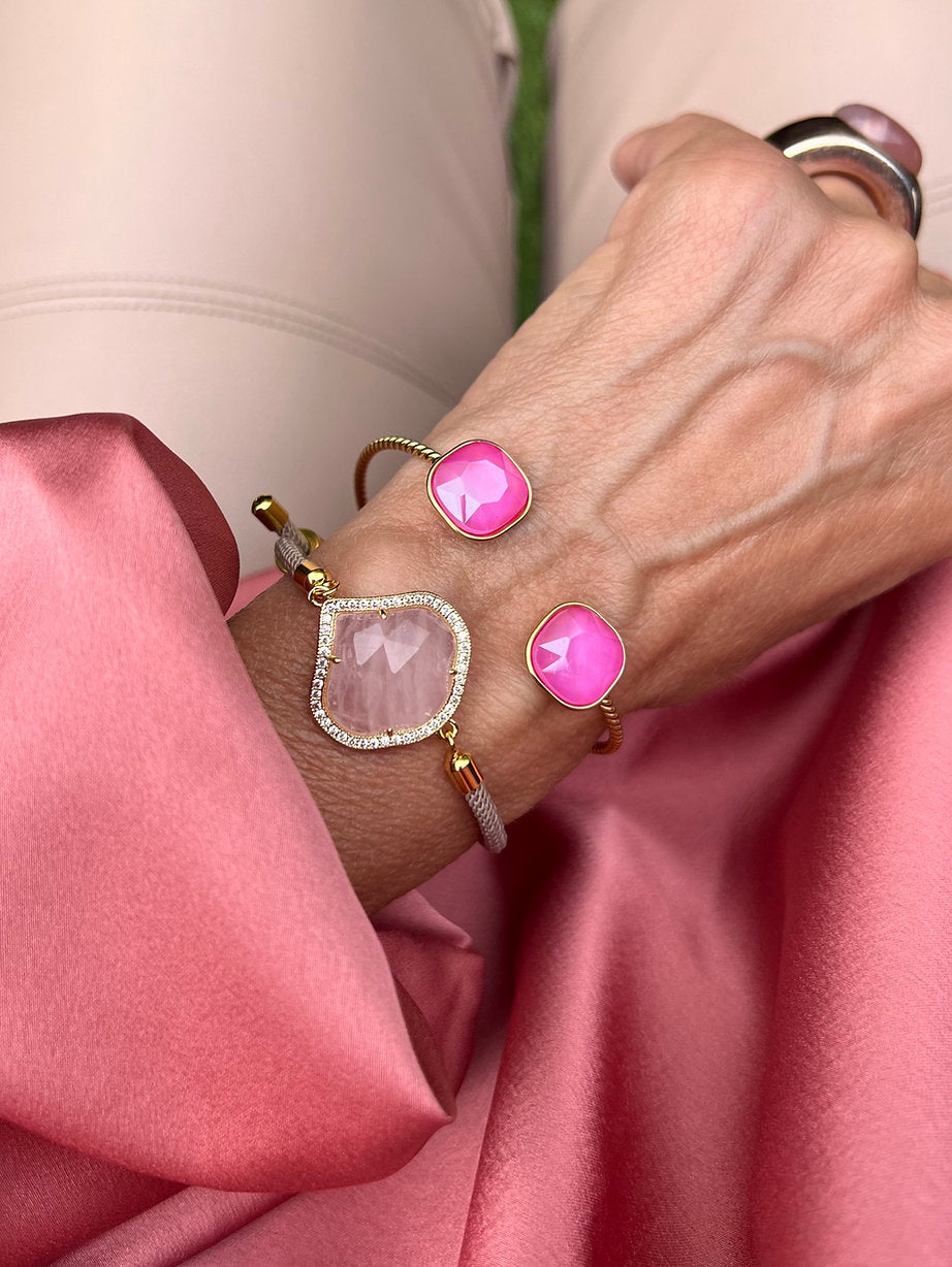 Nahaufnahme eines Handgelenkes mit zwei pinkfarbenen Armbändern, eines mit Cat Eye Stein und roséfarbener facettierter Stein und Zirkonia Rand. die Hand liegt auf einem roséfarbenen Stoff, im Hintergrund eine beigefarbene Hose.