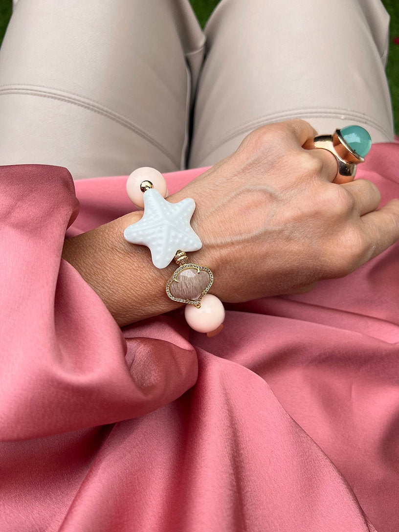 Handgelenk mit rosa Armband aus großen Perlen, verziert mit einem weißen Seestern und einem herzförmigen Kristall.