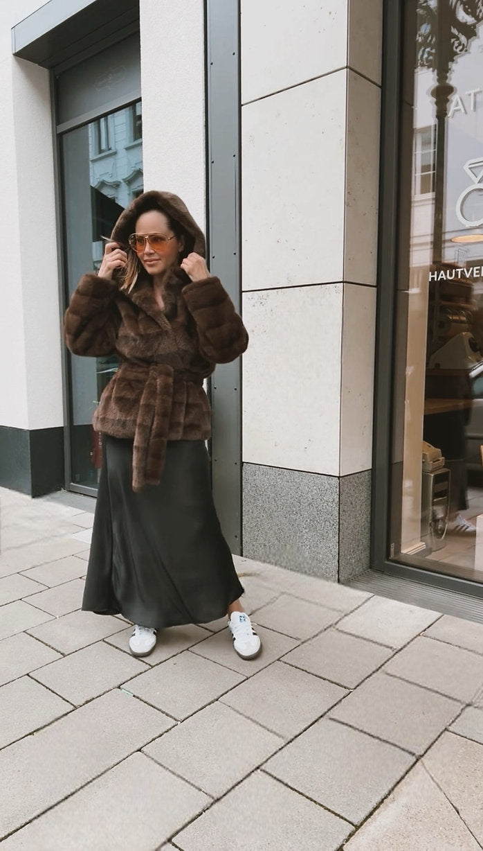 Frau mit Sonnenbrille trägt ein braunes Plüsch Jäckchen mit Bindegürtel und Kapuze, einen weiten schwarzen Rock und weiße Sneaker. Se hält die Kauze mit beiden Händen und steht auf einem Gehweg vor einem Schaufenster.
