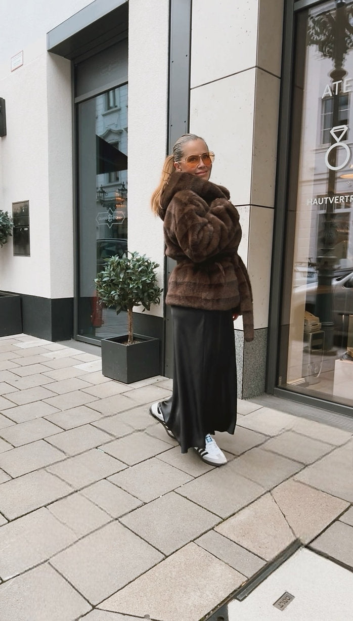 Frau mit Brille trägt ein braunes Plüsch Jäckchen mit Bindegürtel, weiten Rock und weiße Sneaker. Sie steht vor einem Schaufenster in der Stadt.