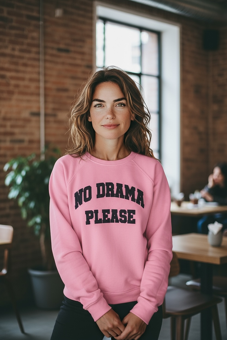 Frau trägt einen pinkfarbenen Pullover mit schwarzen Aufdruck No Drama Please, steht in einem Raum mit Backsteinwand und großen Fenstern.