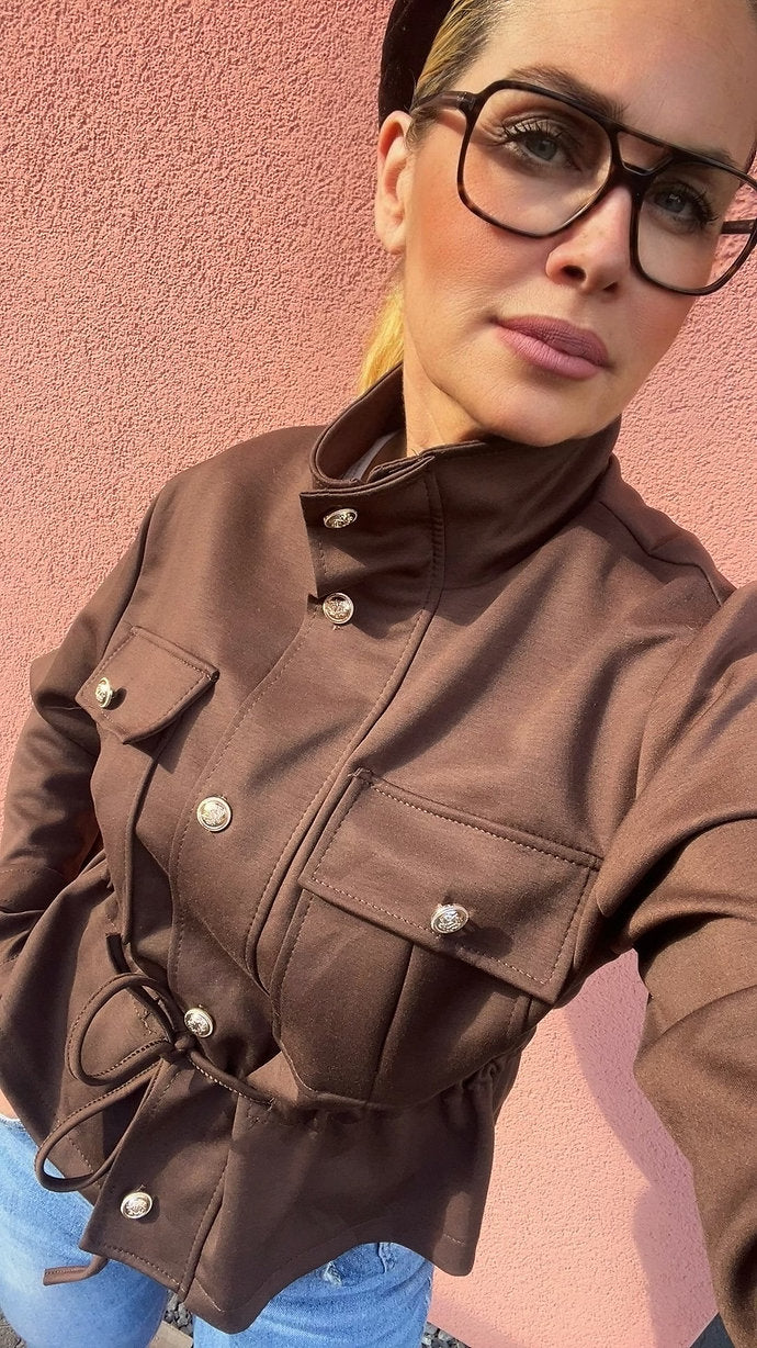 Frau mit Brille und schwarzem Haarreifen steht vor rosa Wand. Braune taillierte Jacke mit einer Jeanshose kombiniert.