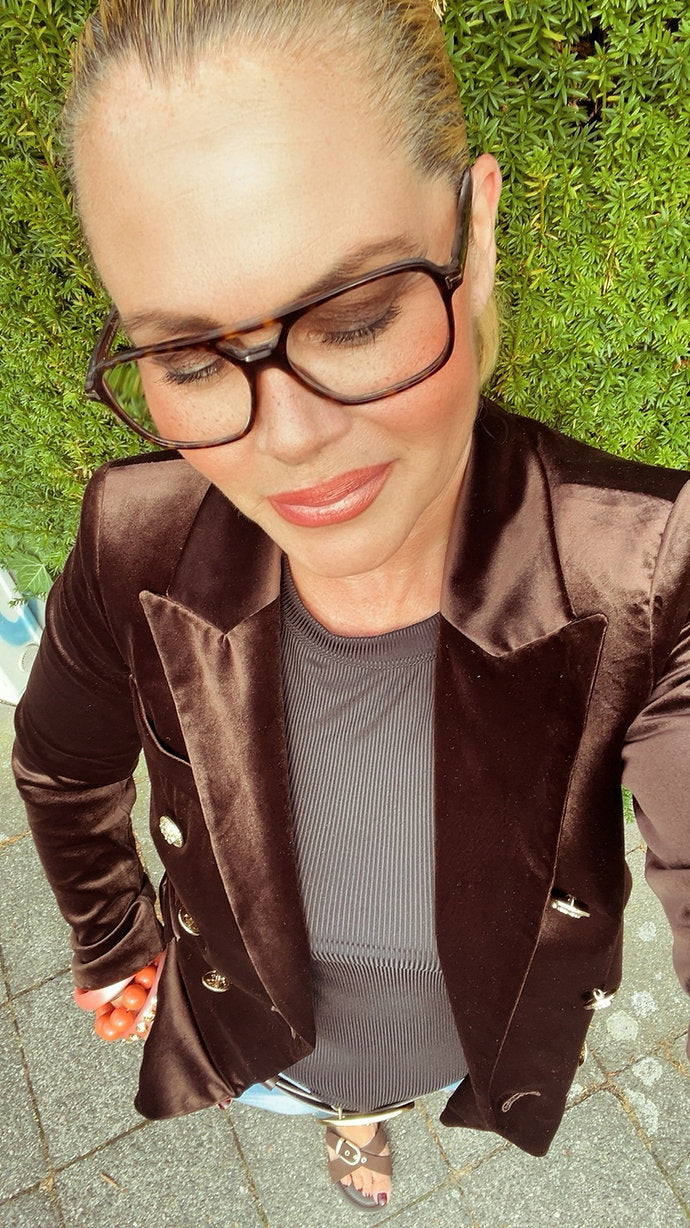 Frau mit zusammengebundenem Haaren trägt einen schobernden Samtblazer mit goldenen Knöpfen, Brille und ein graues Shirt. Selfie von oben, draußen vor grünem Hintergrund.