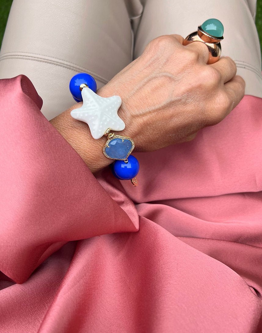 Handgelenk mit blauem Armband aus großen Perlen, verziert mit einem weißen Seestern und einem herzförmigen Kristall.