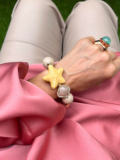 Handgelenk mit sandfarbenen Armband aus großen Perlen, verziert mit einem gelben Seestern und einem herzförmigen Kristall.