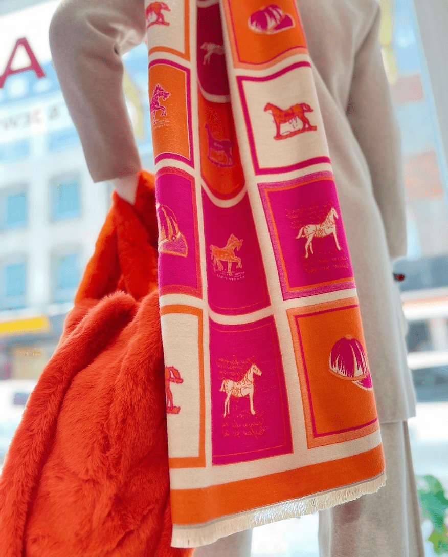 Schaufensterpuppe trägt Schal in pink, Orange und Beige mit Karomuster. Über die Schulter gelegt. Trägt orangefarbene Tasche in der Hand.