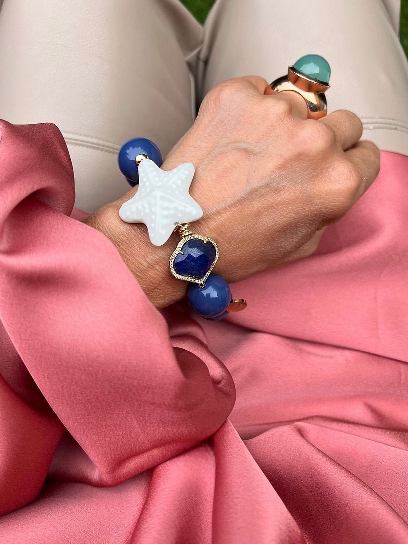 Handgelenk mit blauem Armband aus großen Perlen, verziert mit einem weißen Seestern und einem herzförmigen Kristall.