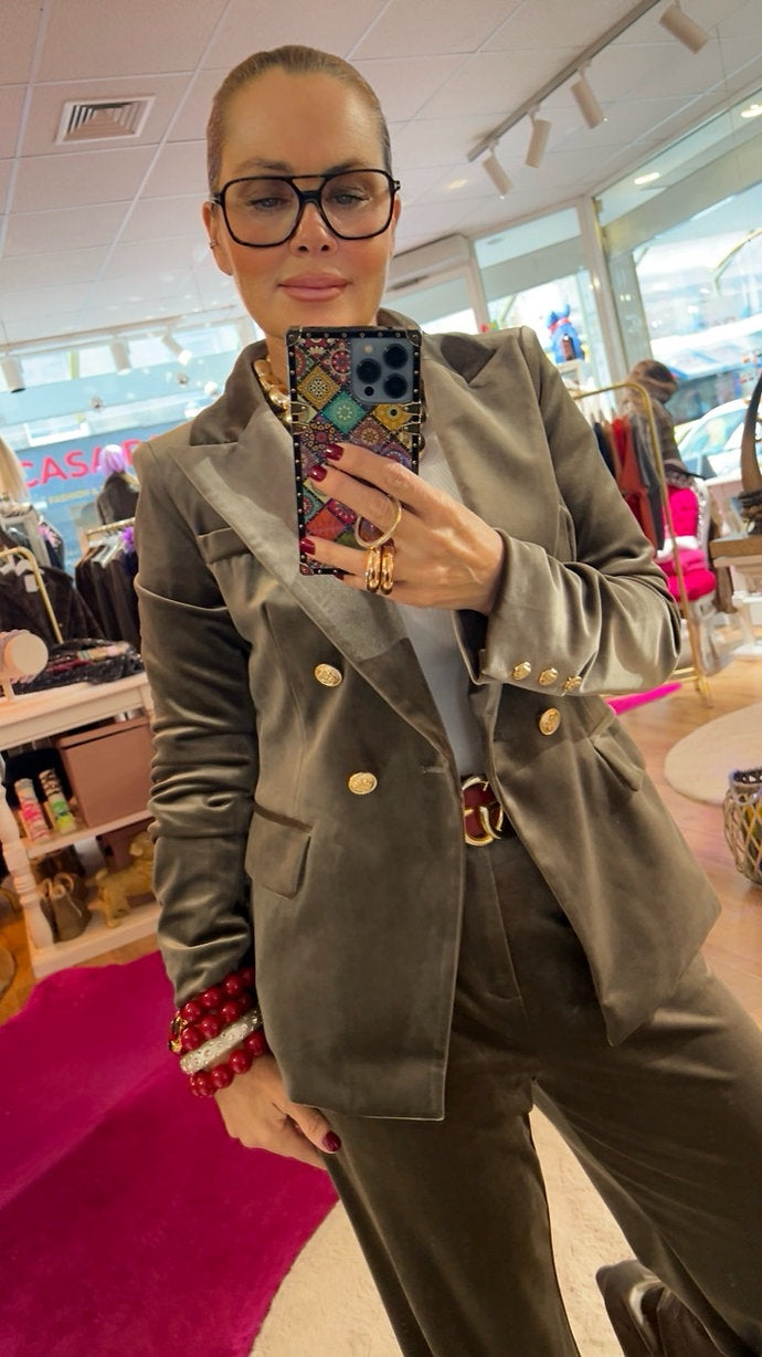 Frau trägt Anzug aus Samt in Taupe mit goldenen Knöpfen. Ein roter Gürtel, Armbänder in Rot. Hält Handy. Fotografiert in einer Boutique.