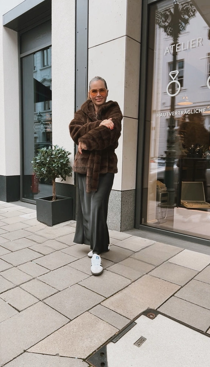 Frau trägt schwarzen Satinrock in A-Linie mit weißen Seakern und brauner Felljacke, fotografiert vor einer Boutique.