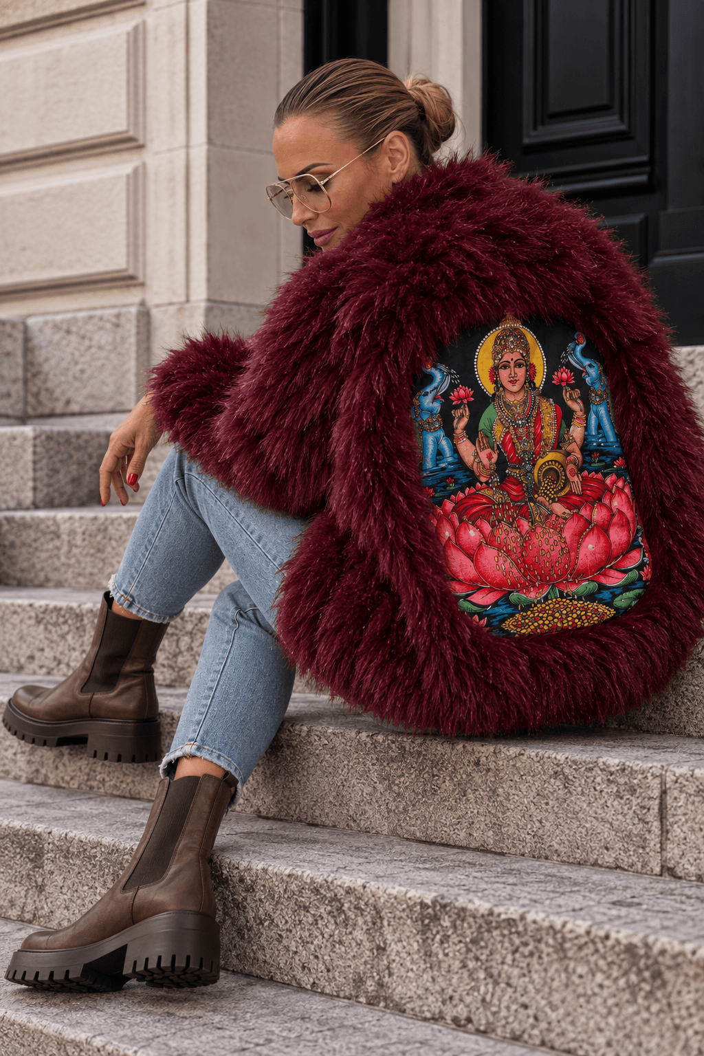 Frau trägt die bordeauxrote CASA Pari Fluffy Jacket mit farbigem Rückenmotiv, Skinny Jeans und braunen Chelsea Boots auf einer Steintreppe.