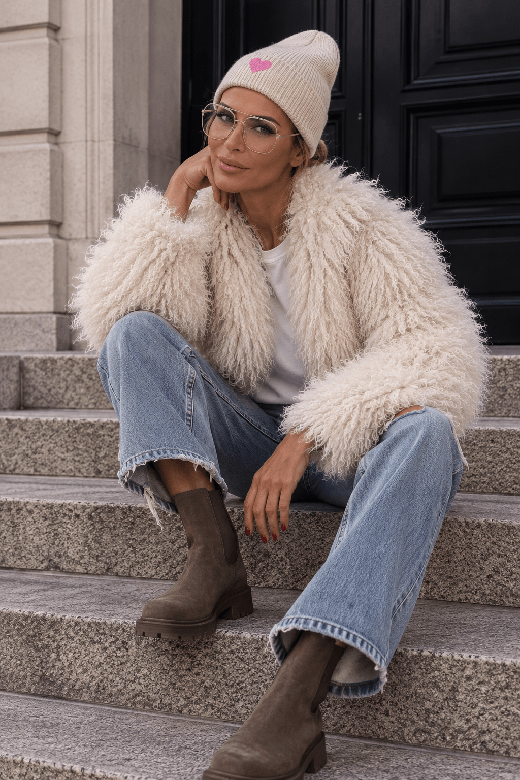 Frau sitzt auf einer Treppe und trägt die cremefarbene CASA Pari Fluffy Jacket zu heller Jeans, braunen Boots und cremefarbener Beanie.