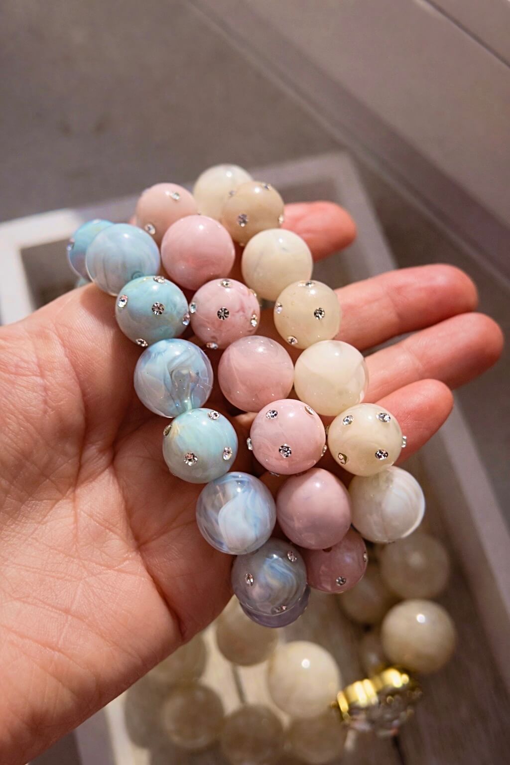 Drei elastische Armbänder mit großen, marmorierten Perlen in Rosé, Blau und Champagner mit kleinen Strasssteinen, auf einer Hand liegend.