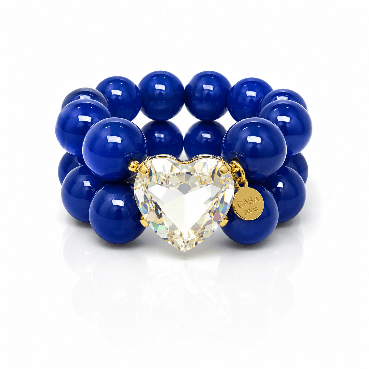 Armband aus glänzenden saphirblauen Resin-Perlen mit großem facettiertem Kristallherz in Champagner-Optik, goldfarbener Fassung und rundem Casapari-Anhänger auf weißem Hintergrund.