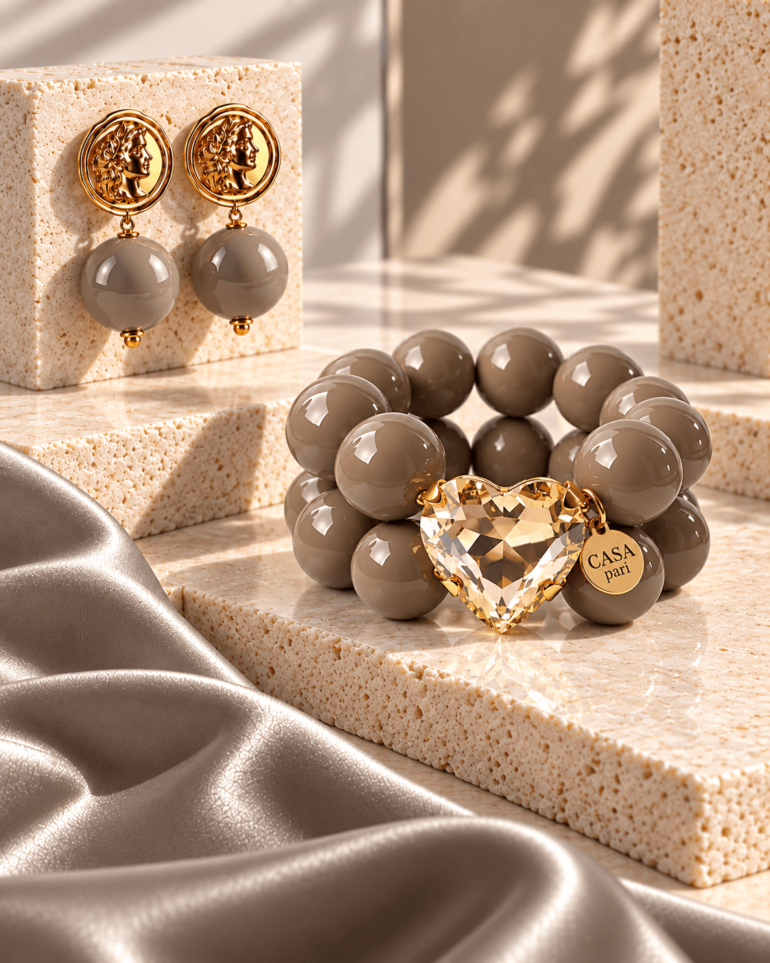 Cashmere-grey Schmuckset von Casapari mit doppelreihigem Perlenarmband, champagnerfarbenem Kristallherz und goldfarbenen Ohrringen auf hellem Travertin.