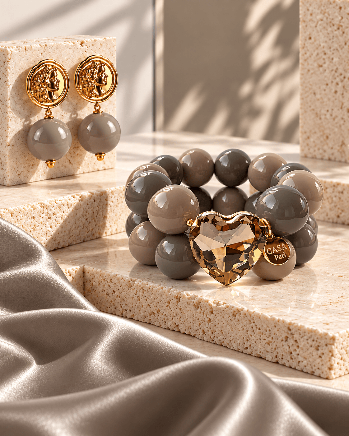 Cashmere-Grey-Mocha Schmuckset von Casapari mit goldfarbenen Ohrsteckern, doppelreihigem Perlenarmband in Grau- und Taupetönen sowie champagnerfarbenem Kristallherz auf hellem Travertin.