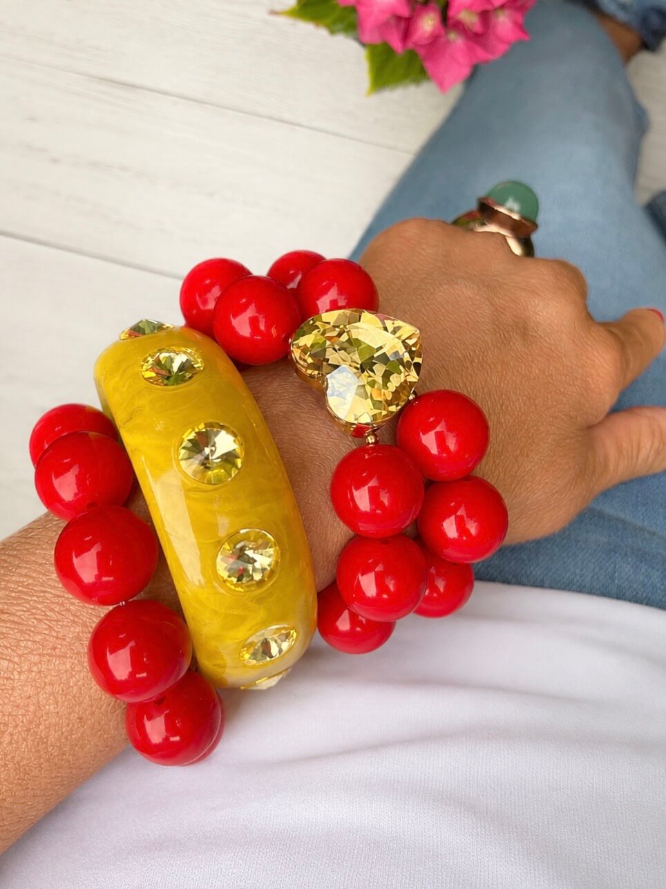 Mehrlagiges Armband-Set mit rotem Crystal-Heart-Armband, gelbem Bangle und weiteren roten Schmuckarmbändern am Handgelenk, kombiniert mit weißem Oberteil und Jeans.
