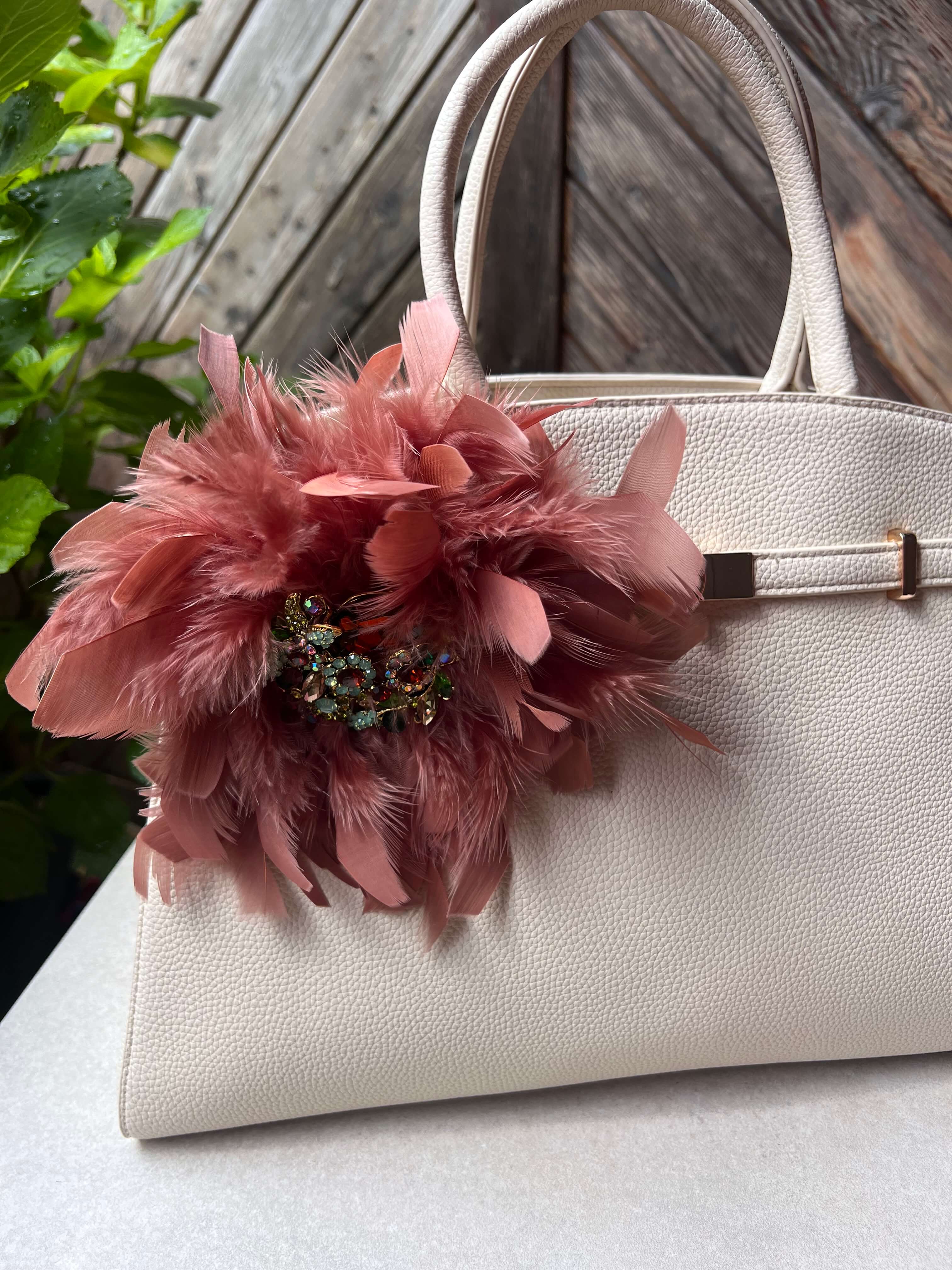 Rosafarbene Federbrosche mit Schmuckelement an einer hellen Handtasche. Die Brosche ist seitlich an der Tasche befestigt und vor einer Holz- und Pflanzenkulisse im Tageslicht fotografiert.