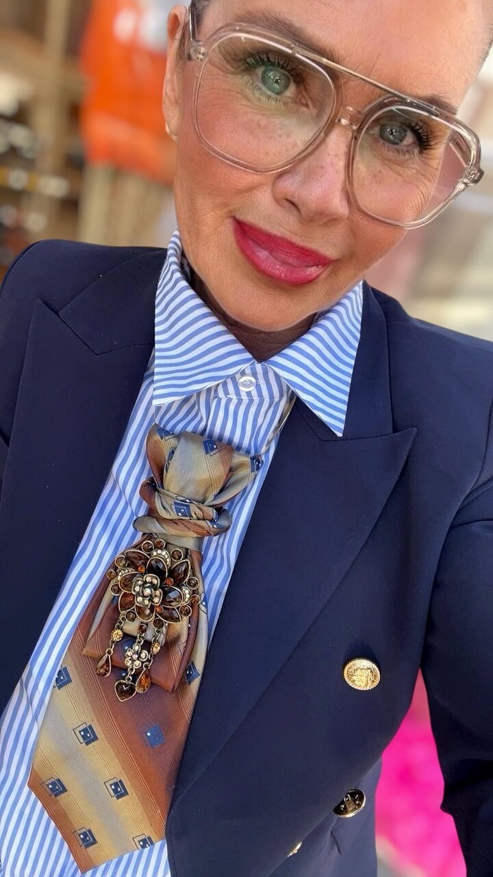 Frau mit transparentem Brillenrahmen trägt dunkelblauen Blazer und blau-weiß gestreifte Bluse, kombiniert mit einer Krawatte in Cognac- und Goldtönen sowie einer dekorativen Brosche mit braunen und goldfarbenen Steinen.