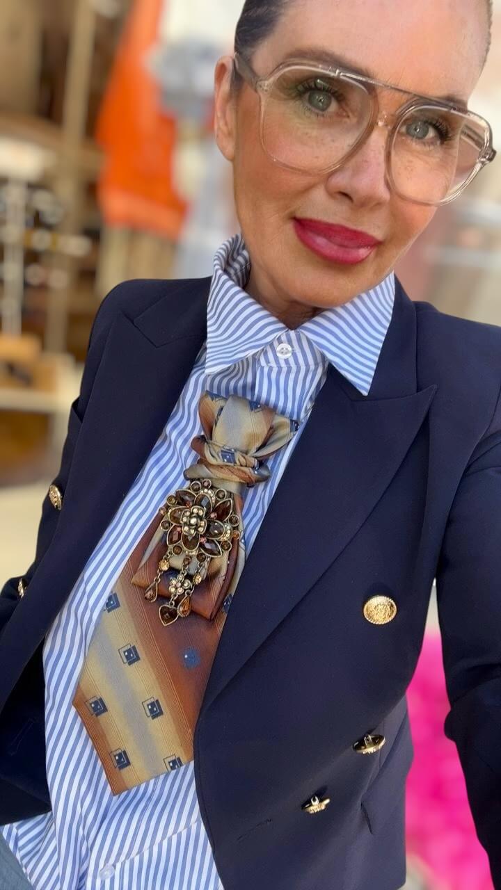 Frau mit transparentem Brillenrahmen trägt dunkelblauen Blazer und blau-weiß gestreifte Bluse, kombiniert mit einer Krawatte in Cognac- und Goldtönen sowie einer dekorativen Brosche mit braunen und goldfarbenen Steinen.