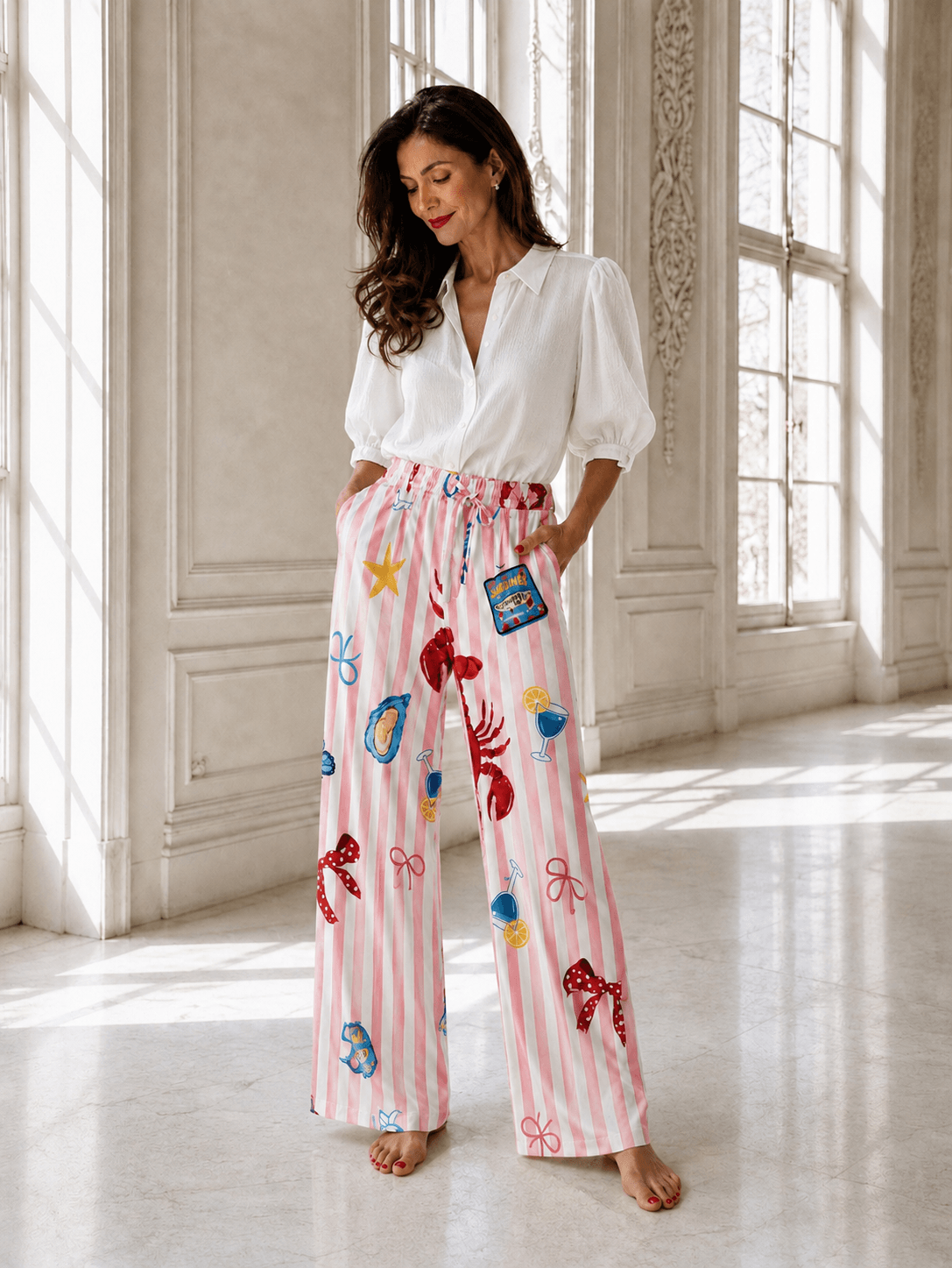 Frau trägt eine rosa-weiß gestreifte Marlene-Hose mit maritimen Motiven wie Hummer, Muscheln und Cocktailgläsern sowie eine weiße Bluse.
