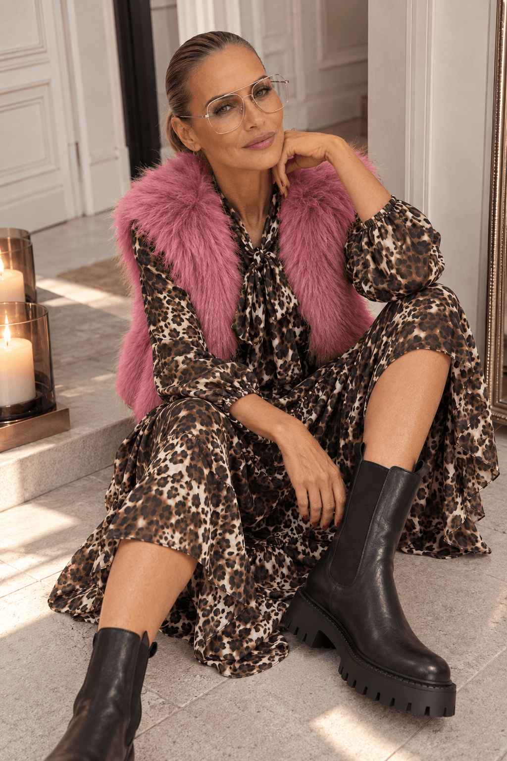 Frau im Leo Bow Dress mit pinker Fake-Fur-Weste und schwarzen Chelsea Boots, lässig sitzend im hellen Innenraum.