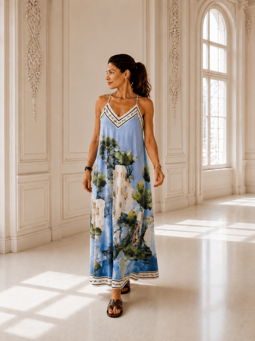 Frau trägt ein bodenlanges Trägerkleid mit grafischem Muster in Blau, Creme und Braun. Das Kleid hat einen fließenden Schnitt und wird in einem hellen Innenraum mit hohen Fenstern präsentiert.