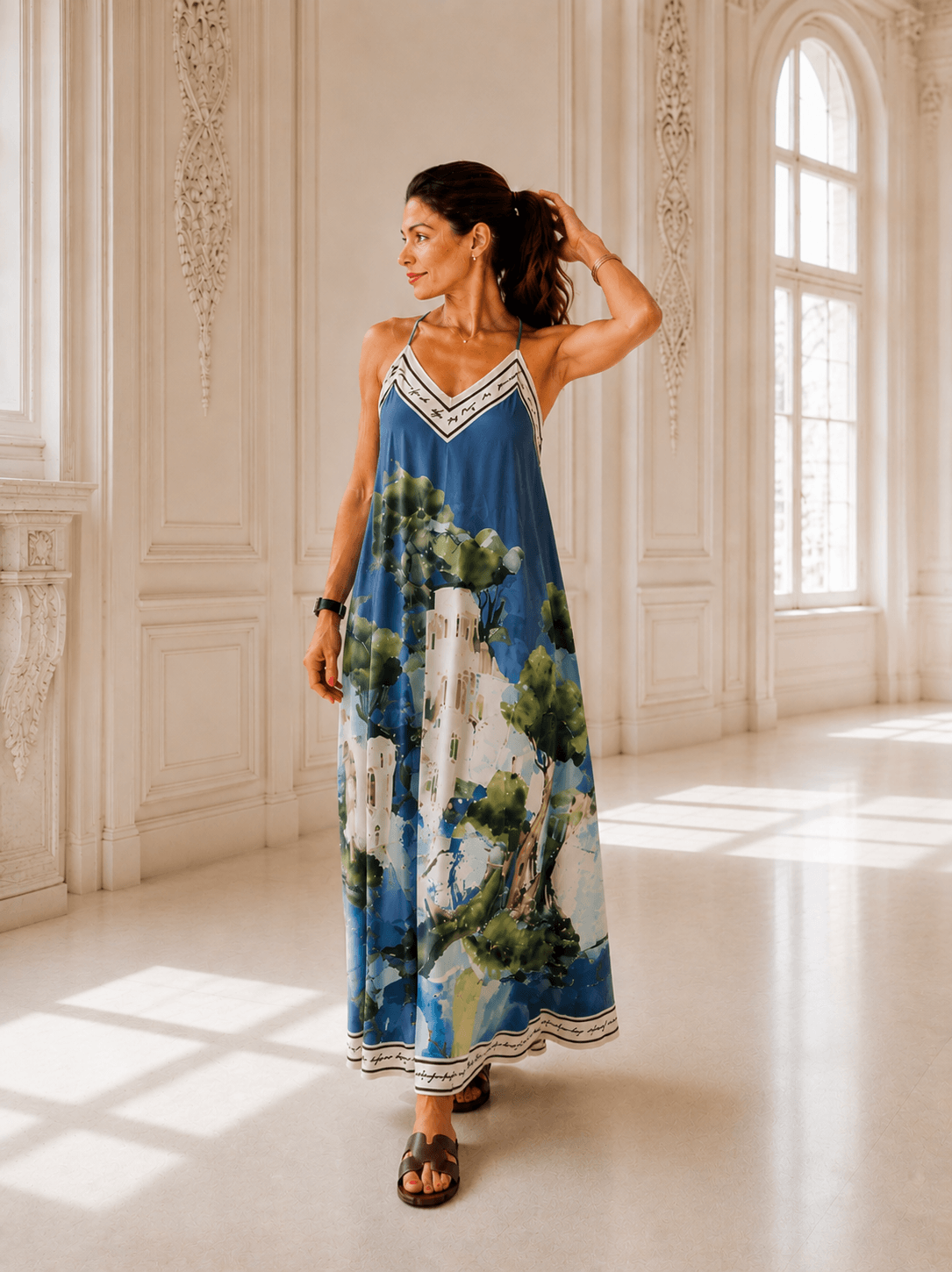 Frau trägt ein bodenlanges Trägerkleid mit grafischem Muster in Blau,Creme und Braun. Das Kleid hat einen fließenden Schnitt und wird in einem hellen Innenraum mit hohen Fenstern präsentiert.