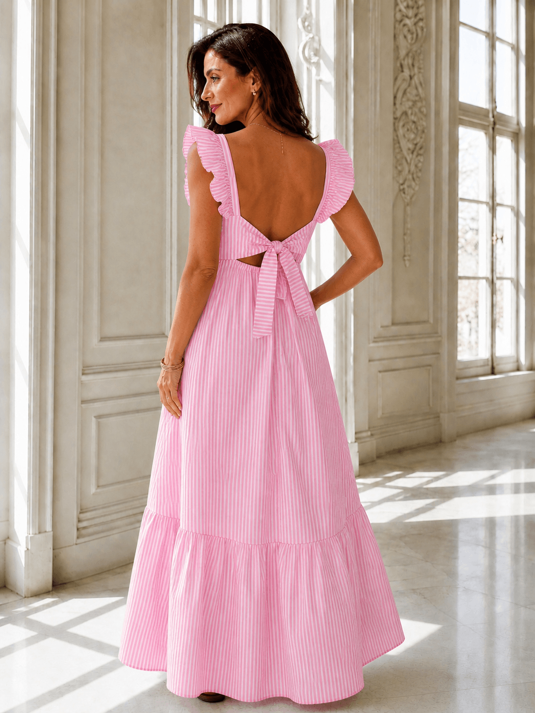 Frau trägt ein rosa-weiß gestreiftes Maxikleid mit Rückenausschnitt und Schleife, steht in lichtdurchflutetem Raum.