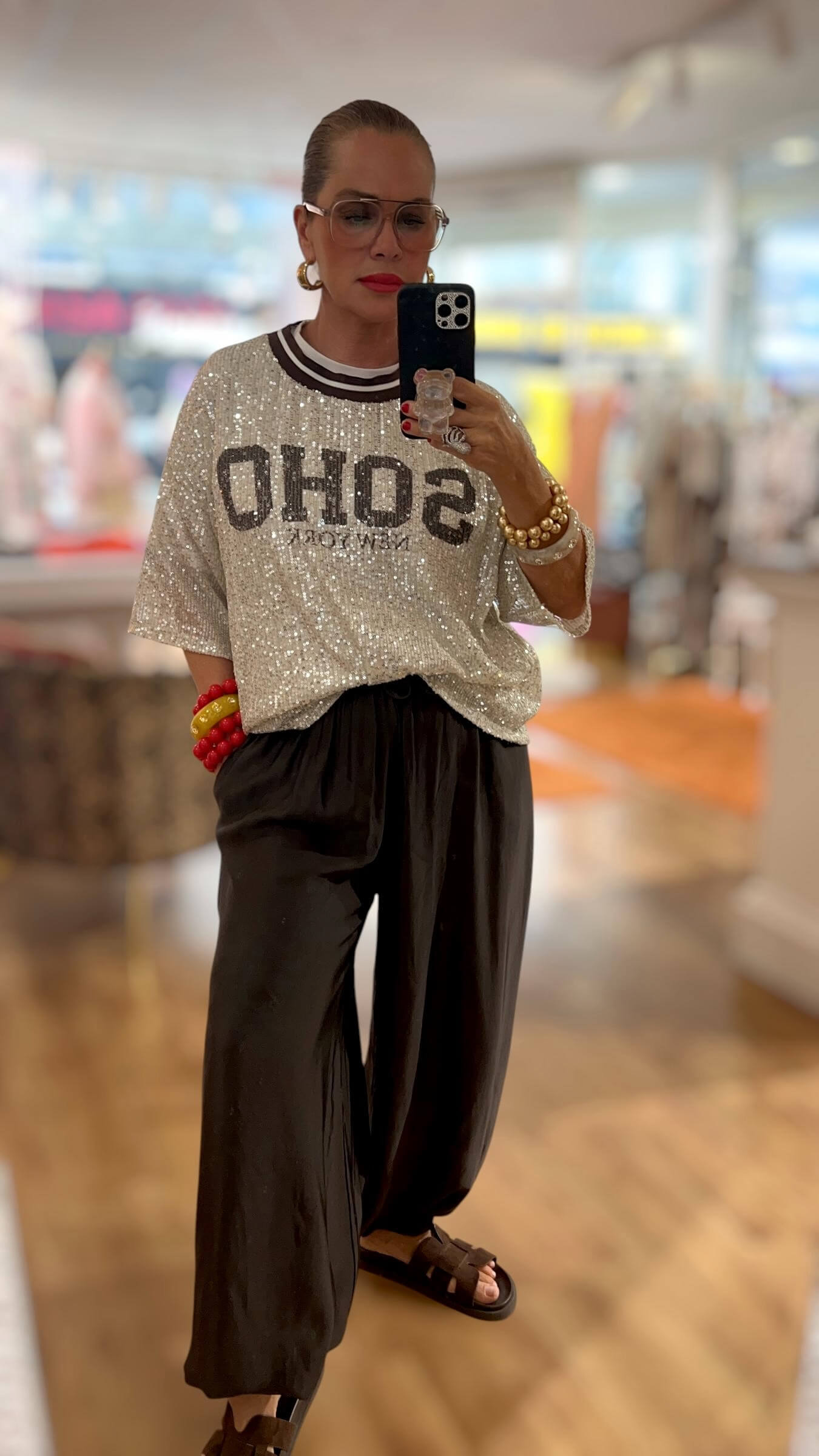 Frau trägt ein silbernes Pailletten-Shirt mit schwarzem Statement-Schriftzug, schwarze weite Hose, braune Sandalen sowie goldene und rote Armreifen in einer Boutique.