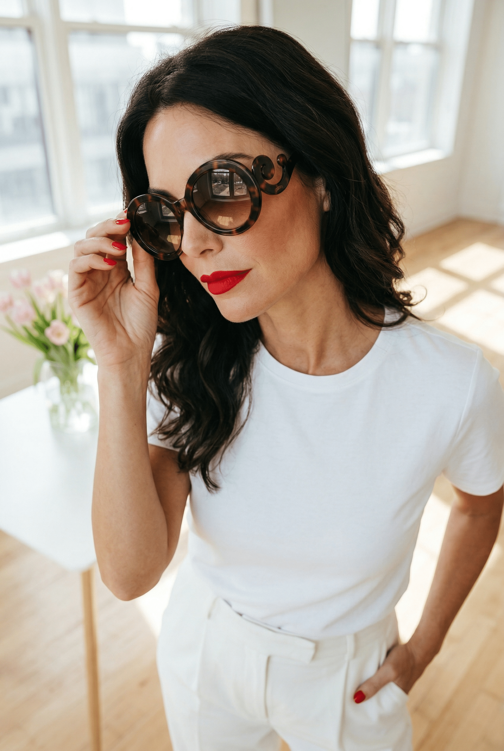 Frau mit dunklen, schulterlangen Haaren trägt eine große runde Sonnenbrille in Braun mit auffälligem Bügeldetail; sie steht in einem hellen Raum am Fenster, trägt ein weißes Outfit und hält die Brille leicht am Rand fest.