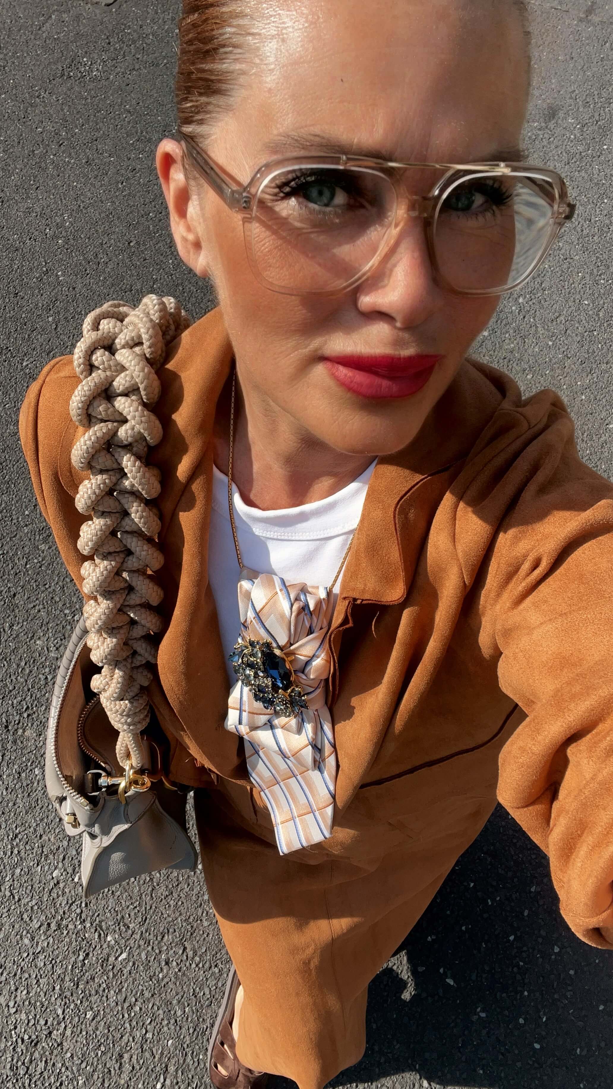 Frau mit camelfarbenem Blazer und weißem Oberteil trägt eine beige karierte Statement-Krawatte mit Schmuckbrosche und transparenter Brille.