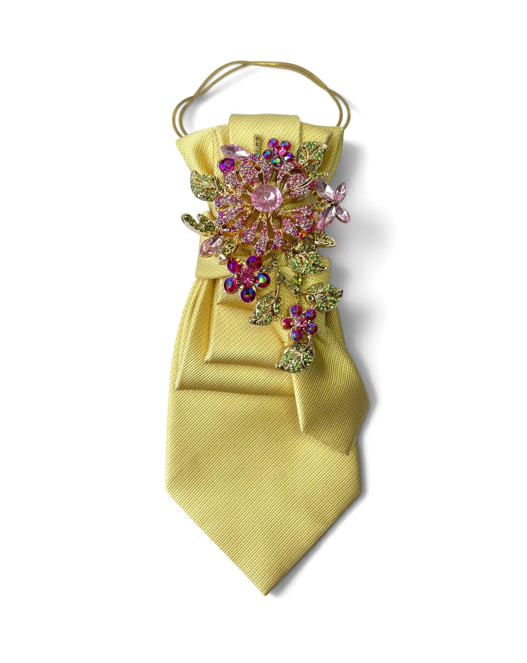 Pastellgelbe Statement-Krawatte von Casapari mit floraler Schmuckbrosche in Rosa, Grün und Gold sowie goldfarbener Kette.