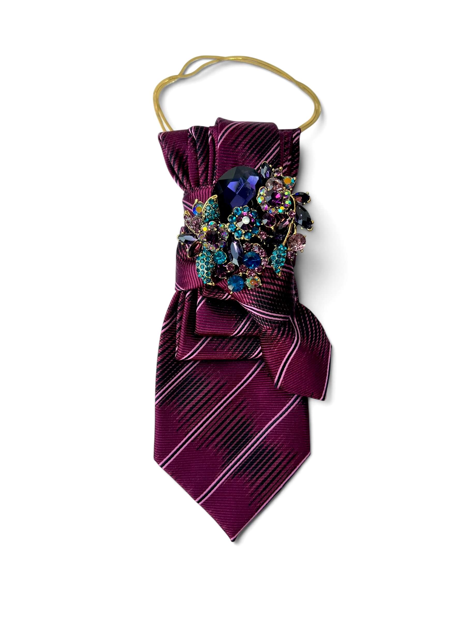 Bordeauxfarbene Statement-Krawatte von Casapari mit funkelnder Schmuckbrosche in Blau, Türkis und Violett sowie goldfarbener Kette.