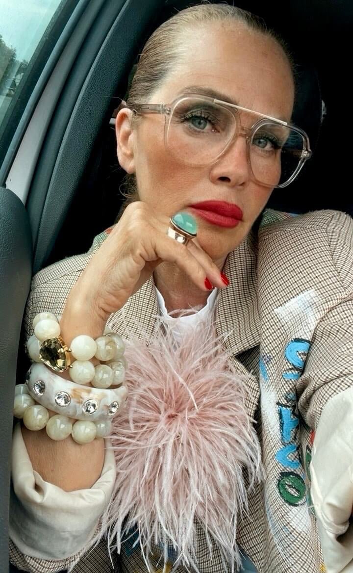 Frau mit transparenter Brille, roten Lippen und gemustertem Blazer sitzt im Auto und präsentiert mehrere helle Armreifen, einen Ring mit türkisfarbenem Stein und eine rosafarbene Federbrosche.