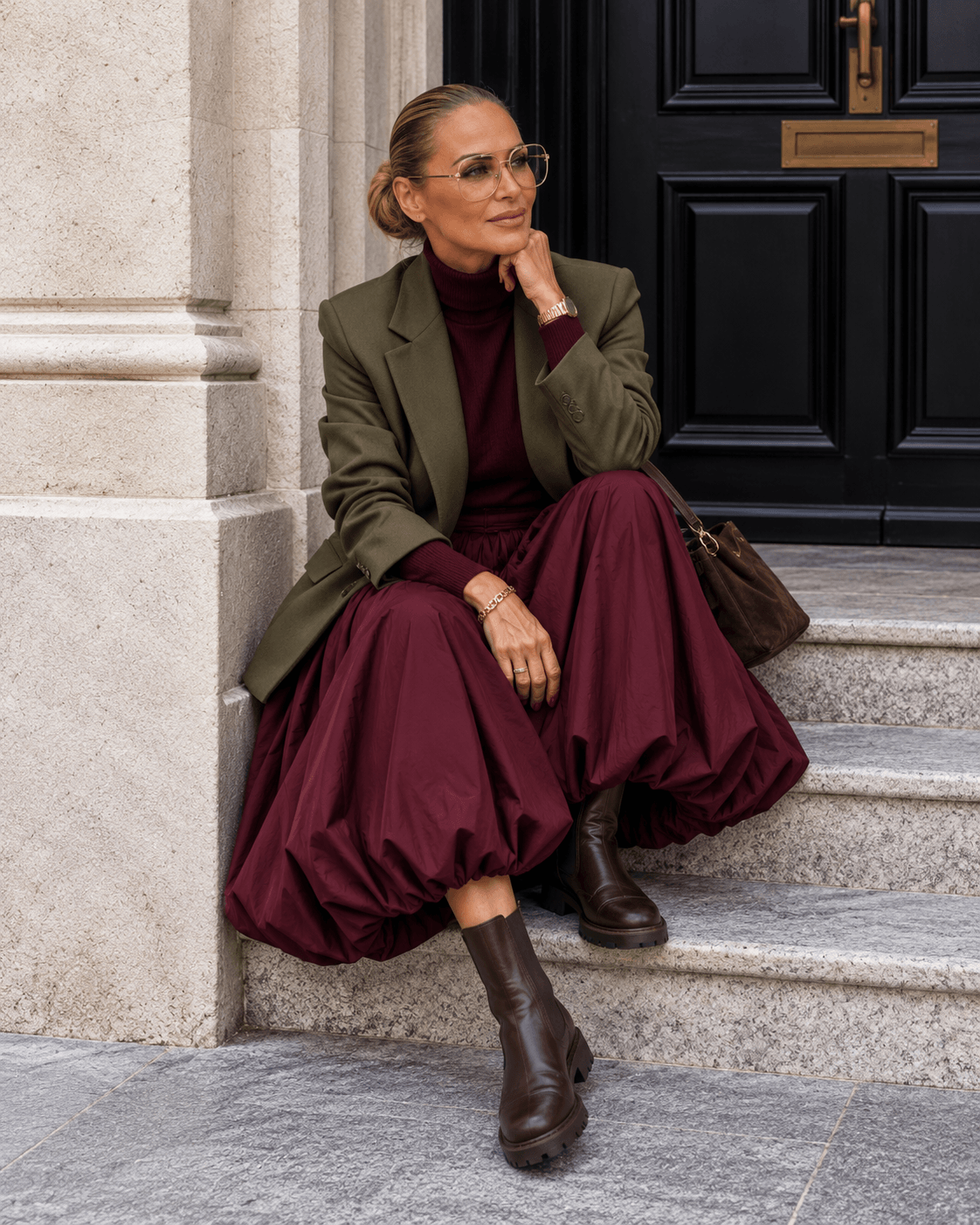 Frau mit burgundyfarbenem Ballonrock, burgundy Rollkragenpullover, olivgrünem Blazer und braunen Chelsea Boots auf einer Steintreppe.