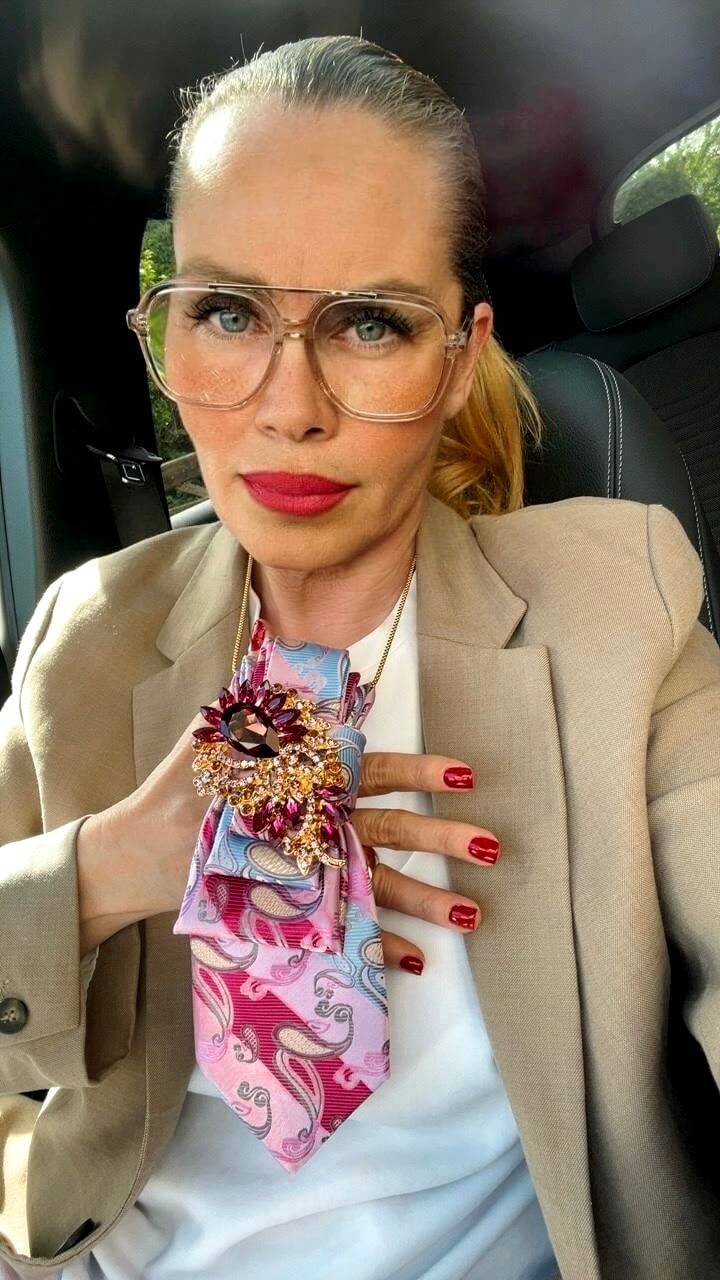 Frau trägt weißen Look mit fliederfarbenem Blazer und eine rosa gemusterte Vintage-Krawatte mit großer Schmuckbrosche in Pink- und Goldtönen als Styling-Detail.