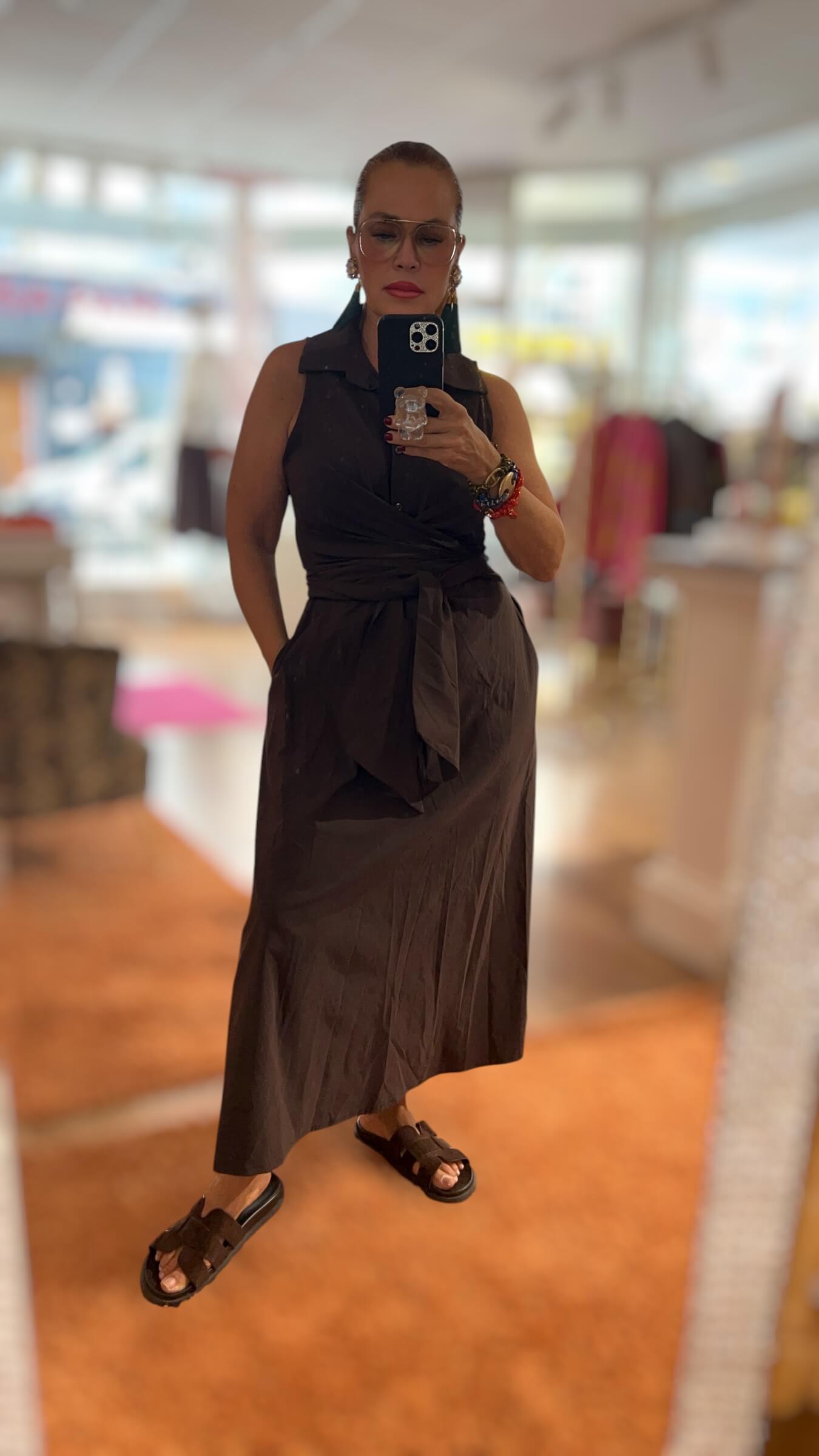 Frau trägt ein ärmelloses Wickelkleid in Espresso-Schwarz mit Hemdkragen und Bindegürtel. Das knöchellange Kleid wird mit dunklen Sandalen, auffälligen Ohrhängern und mehreren Armbändern kombiniert. Fotografiert in einer hellen Boutique.