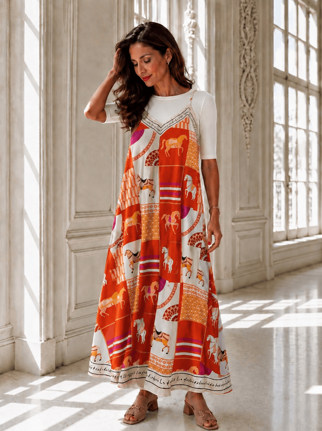 Frau trägt ein orange-weiß gemustertes Maxikleid mit Pferdemotiven über einem weißen, eng anliegenden Halbarmshirt in einem hellen Innenraum mit großen Fenstern.