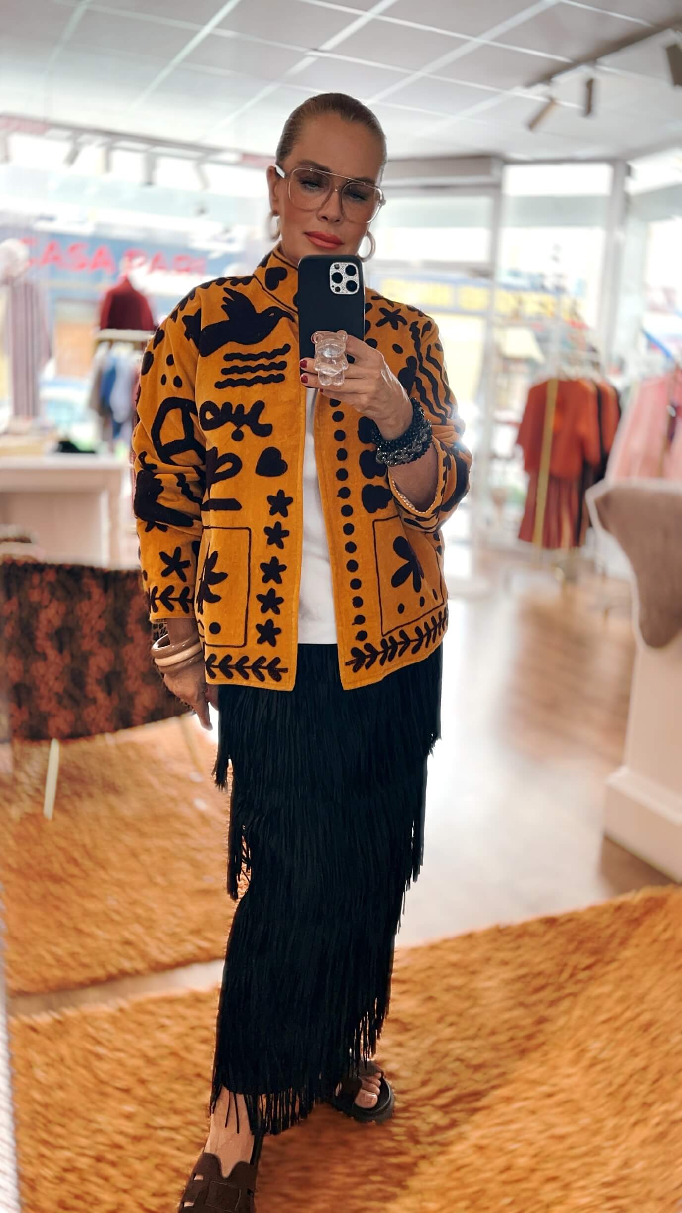 Frau trägt die Embroidered Couture Jacke Golden Saffron mit schwarzer Stickerei über einem weißen Oberteil und einem schwarzen Fransenrock in einer hellen Boutique.