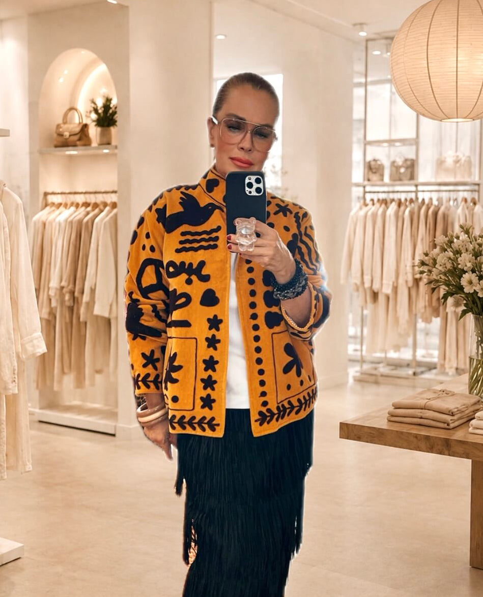 Frau fotografiert sich in einer hellen Boutique. Sie trägt eine ockerfarbene, grafisch bestickte Embroidered Couture Jacke, ein weißes Oberteil und einen schwarzen Fransenrock.