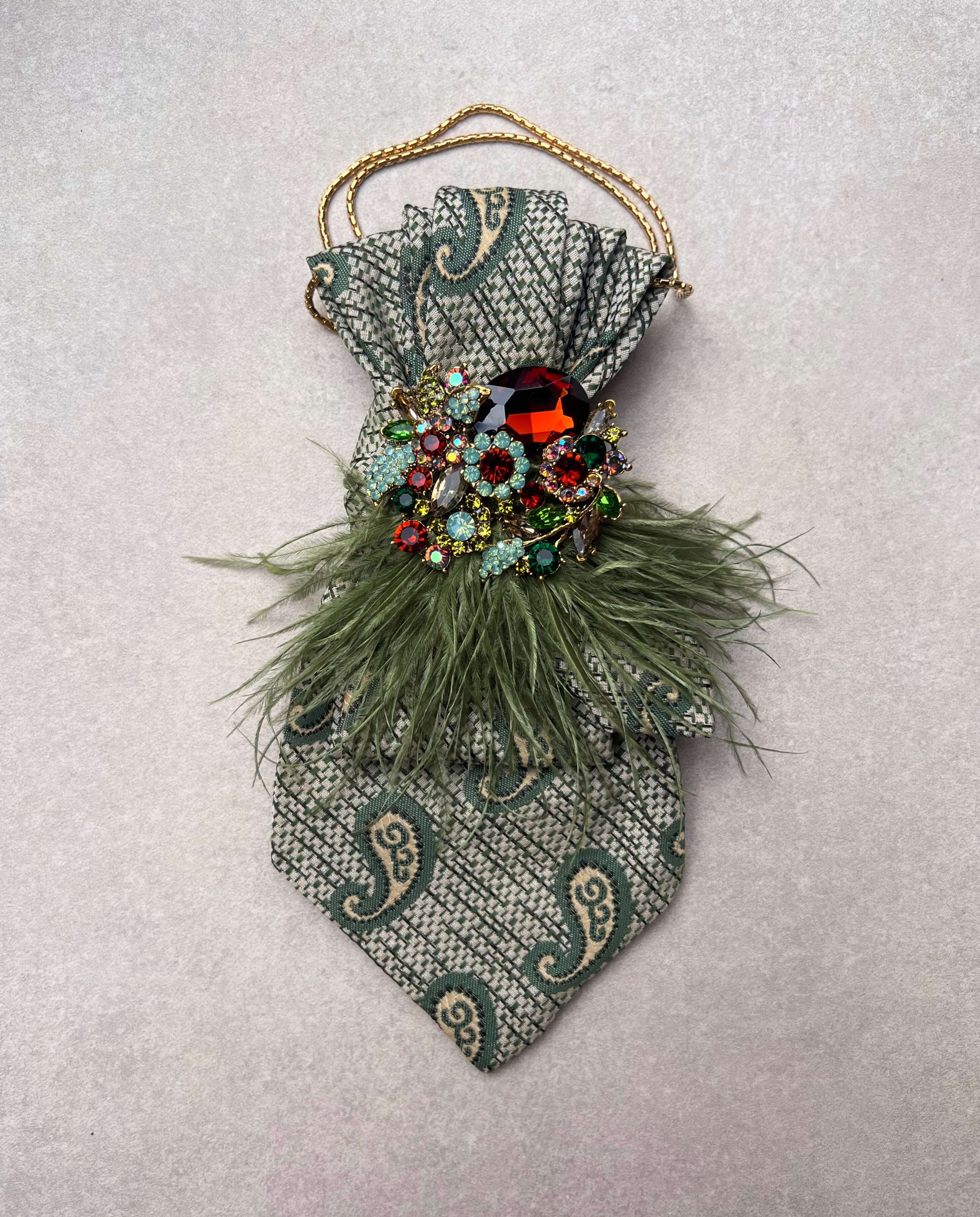 Vintage-Krawatte in Salbeigrün mit Paisley-Muster, grünen Federdetails, goldfarbener Kette und großer Schmuckbrosche mit mehrfarbigen Kristallen auf hellem Hintergrund.