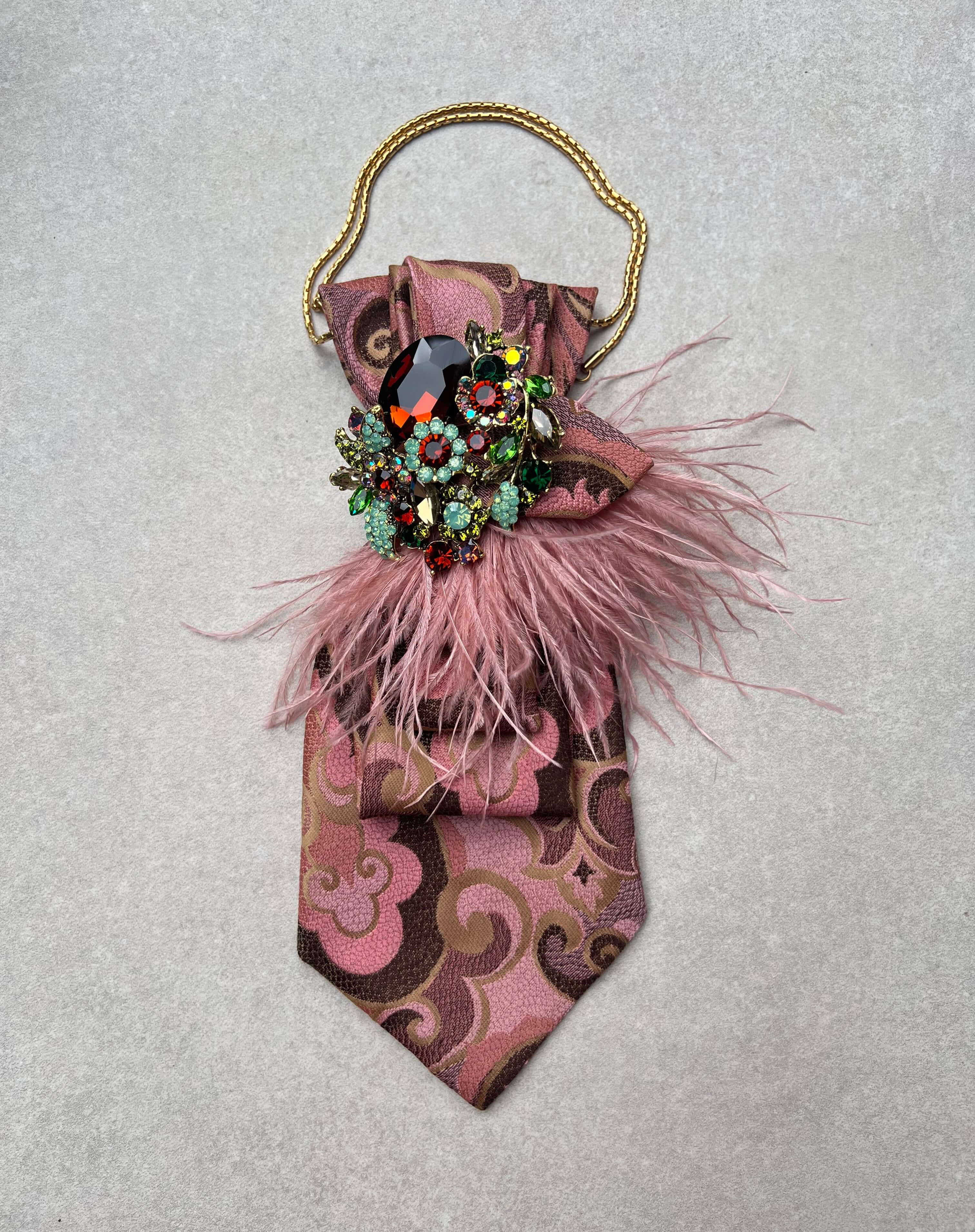 Vintage-Krawatte in Rosé und Braun mit Paisley-Muster, rosafarbenen Federdetails, goldfarbener Kette und großer Schmuckbrosche mit mehrfarbigen Kristallen auf hellem Hintergrund.